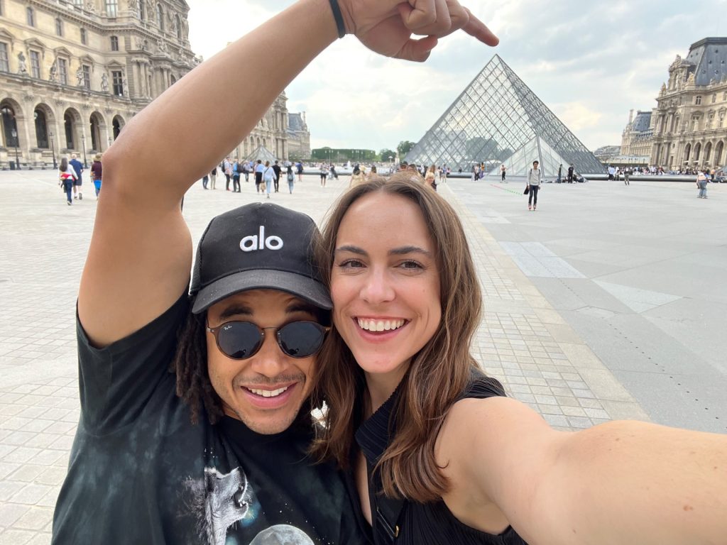 A smiling couple takes a selfie in front of the Louvre Pyramid in Paris. The man wears sunglasses, a black "alo" cap, and a graphic tee, while the woman has long brown hair and a black top—capturing their special spotlight moment.