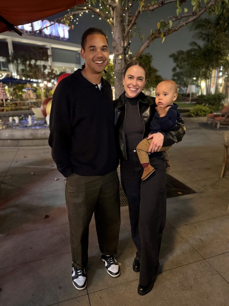 A smiling man and woman stand together outdoors at night, with Megan Spire holding a baby. Festive lights and trees glow in the background, putting a spotlight on their joyful moment in this lively scene.