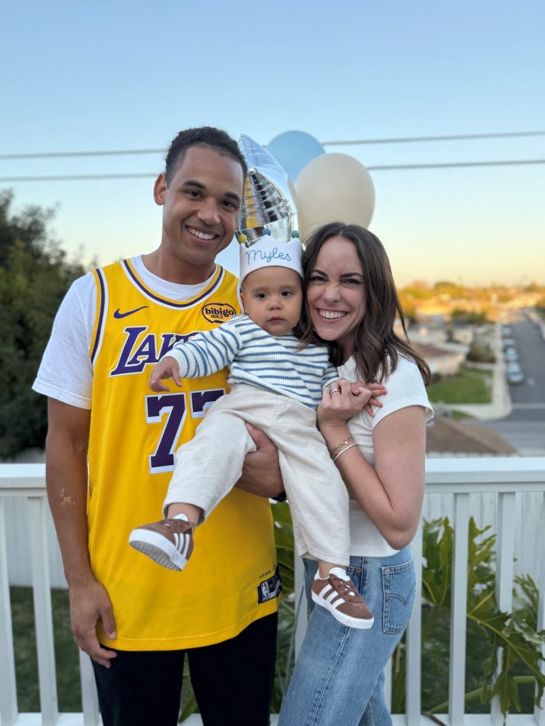 A smiling family poses outdoors: Megan Spire in a yellow Lakers jersey, another adult in a white t-shirt holding baby Myles—wearing a striped shirt and birthday hat. Balloons float nearby, adding flair to the Vendor Advisory-themed celebration.