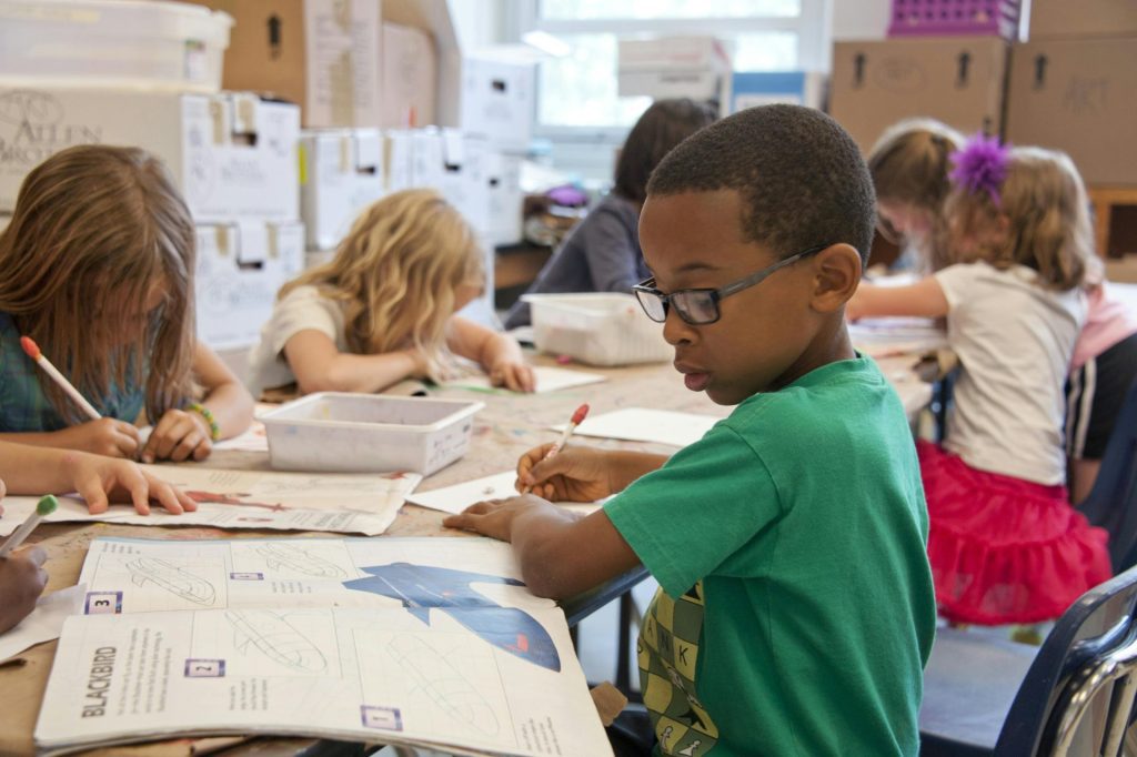 A group of young children sit at a classroom table, drawing and coloring in workbooks, with art supplies scattered around. The focus is on a boy in glasses and a green shirt, engaged in an education project that promotes greener schools.