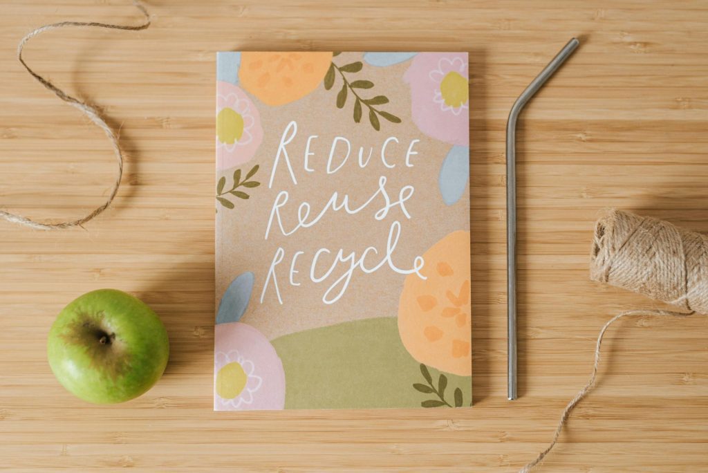 A notebook with "Reduce Reuse Recycle" on the cover is placed on a wooden surface, surrounded by a green apple, a metal straw, twine, and string—promoting Greener Schools and sustainable solutions for education.