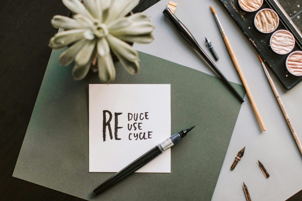 A flat lay of art supplies, including paintbrushes, calligraphy nibs, watercolor paints, and a potted plant, surrounds a card reading "REDUCE REUSE RECYCLE"—perfect inspiration for education on sustainable solutions.