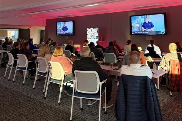 A group of people sit on white chairs in a modern conference room at ASB, watching a 2025 virtual award ceremony unfold on two large screens. The room is lit with natural light and soft pink accent lighting.
