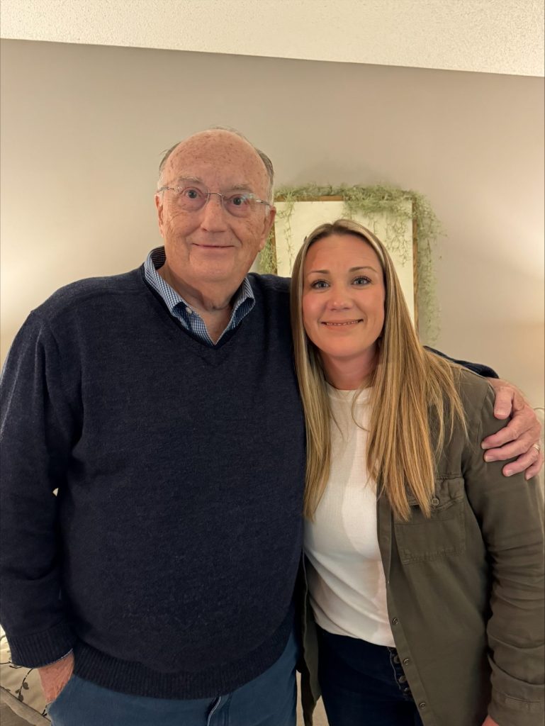 Dennis Rynearson, in a navy sweater and glasses, smiles with his arm around Jennifer Quinnell, who has long blonde hair and a green jacket; both stand indoors against a beige wall—capturing a warm Family Matters moment.