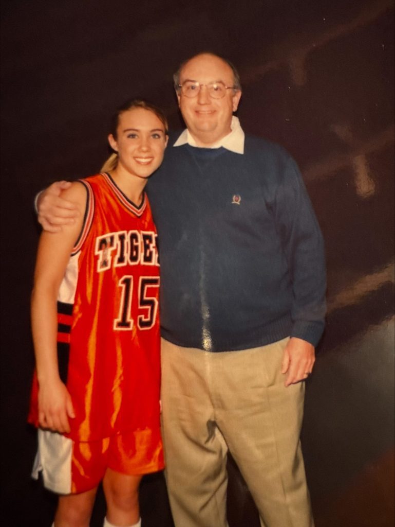 Jennifer Quinnell, in her orange "Tigers" basketball uniform (#15), stands smiling beside Dennis Rynearson—both exuding pride. Against a dark background, this moment beautifully captures the spirit of Family Matters.