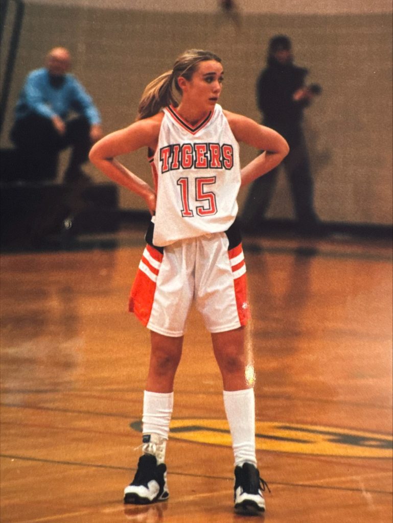 Jennifer Quinnell, wearing a white Tigers jersey with the number 15, stands on the gym court with her hands on her hips, looking to the side as spectators and blurred figures fill the background.