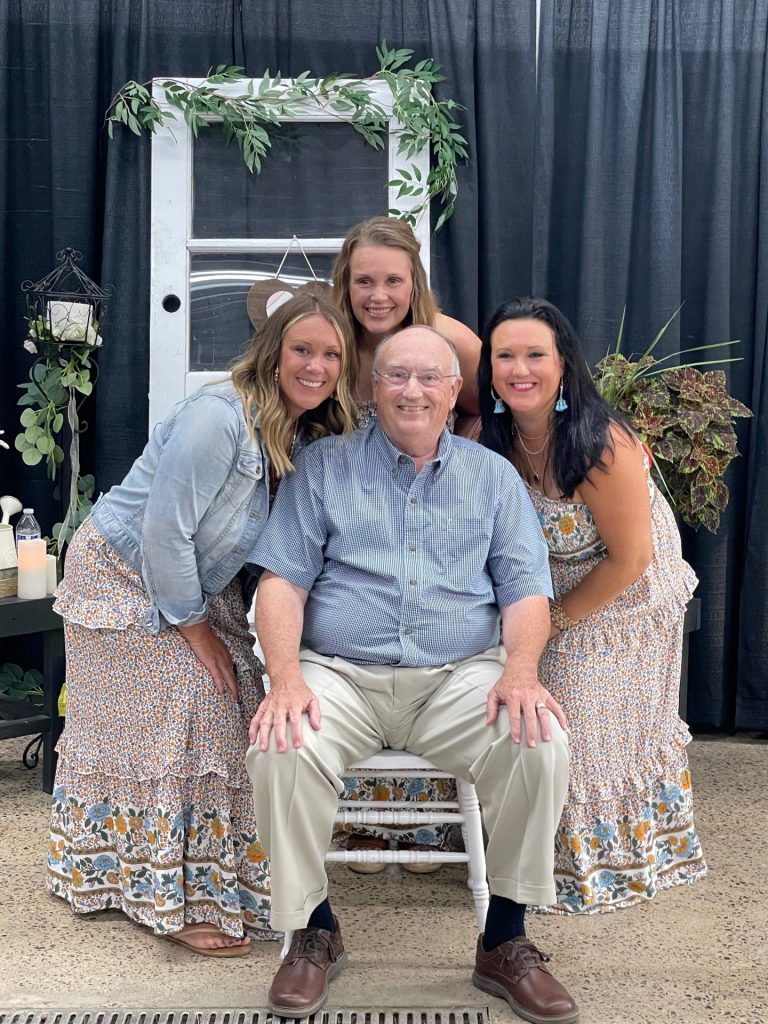Three women in matching floral dresses pose smiling around Dennis Rynearson, who is seated on a white chair. Indoors, with a decorative backdrop of plants and a vintage door, this moment captures the warmth that Family Matters most.