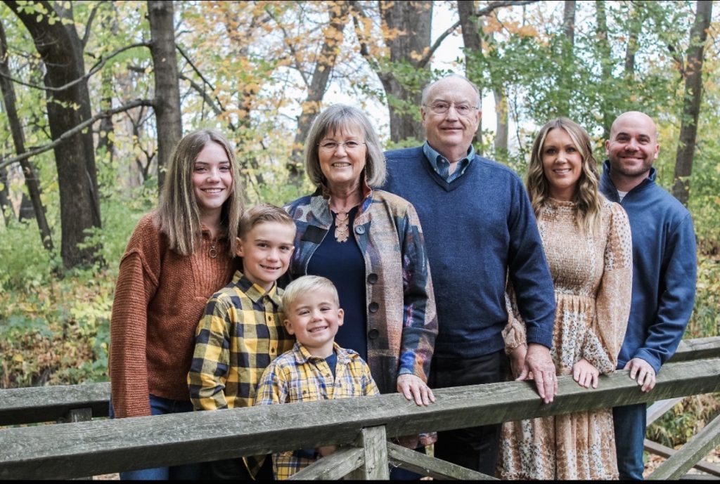 Jennifer Quinnell and Dennis Rynearson, along with their loved ones, stand together on a wooden bridge in a forested area with autumn foliage, smiling and posing for a Family Matters-inspired photo.