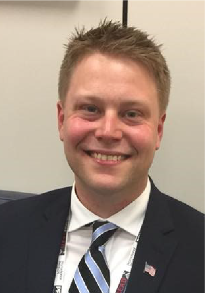 A man in a suit and striped tie smiles at the camera, celebrating his role among esteemed ASB Advisory Board Members. He has short, light brown hair, a U.S. flag lapel pin, and stands against neutral-colored indoor walls.