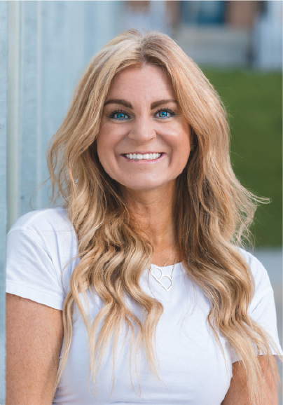 A woman with long wavy blonde hair and bright blue eyes smiles at the camera. She is wearing a white shirt and a heart-shaped necklace, standing outdoors with a blurred green background, embodying the approachable spirit of our Advisory Board Members.