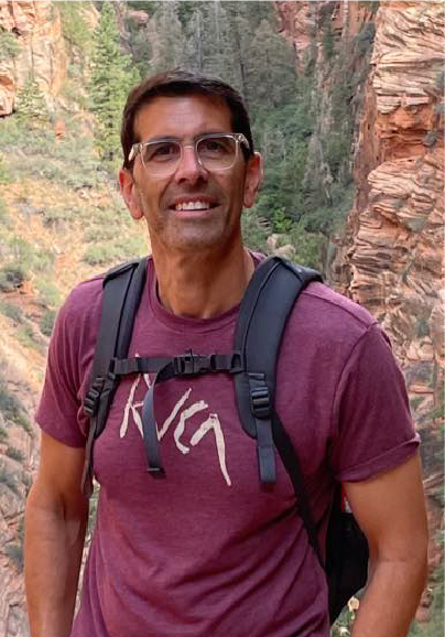 A man wearing glasses, a maroon T-shirt, and a black backpack smiles while standing outdoors in a rocky canyon as he celebrates with fellow ASB Advisory Board Members, surrounded by trees in the background.