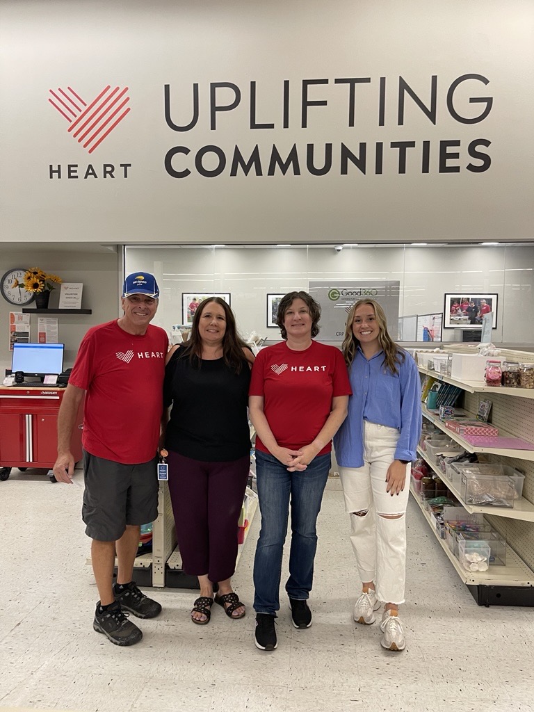 Four people stand smiling inside a store, under a sign reading “Heart Uplifting Communities.” Two wear matching red shirts with a heart logo, showcasing the Non-Profit HEART partnership with American Solutions for Business. Shelves and photos fill the background.