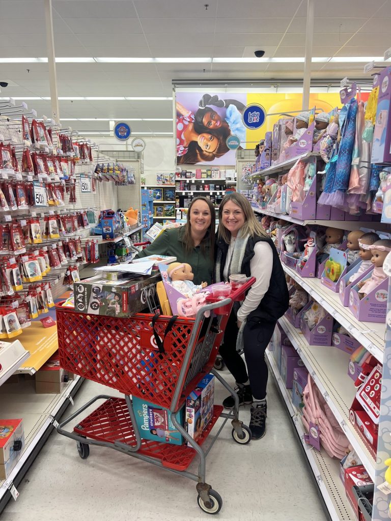Two women smile next to a red shopping cart filled with toys in a store aisle lined with dolls, coloring books, and children's toys, gathering gifts for ASB's annual Adopt-A-Family initiative. Bright store lighting and colorful displays fill the background.