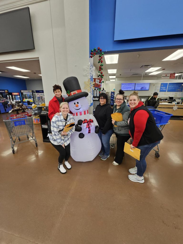 Five people pose and smile around a large inflatable snowman inside a store during the ASB annual fundraiser. Shopping carts and a counter are visible in the background, with some people holding yellow envelopes for Adopt-A-Family donations.