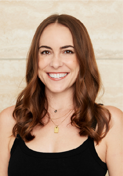 A woman with long, wavy brown hair smiles at the camera, celebrating her role among ASB Advisory Board Members. She wears a black sleeveless top and layered gold necklaces, set against a light beige stone wall background.