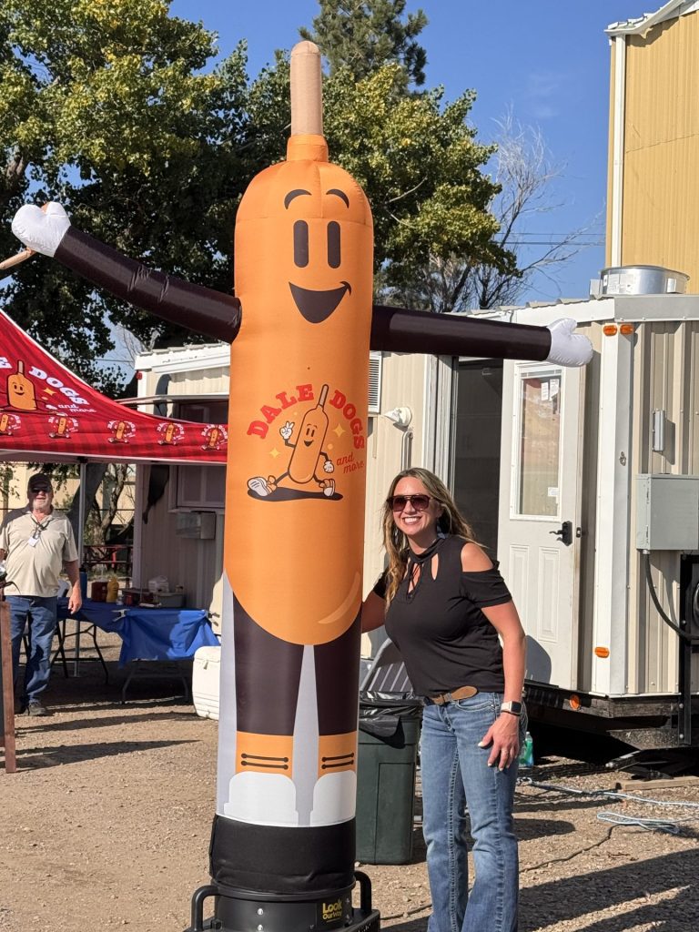 A woman wearing sunglasses and a black shirt smiles while standing next to a tall, inflatable hot dog mascot with raised arms and a happy face at an outdoor event featuring vibrant custom signage.