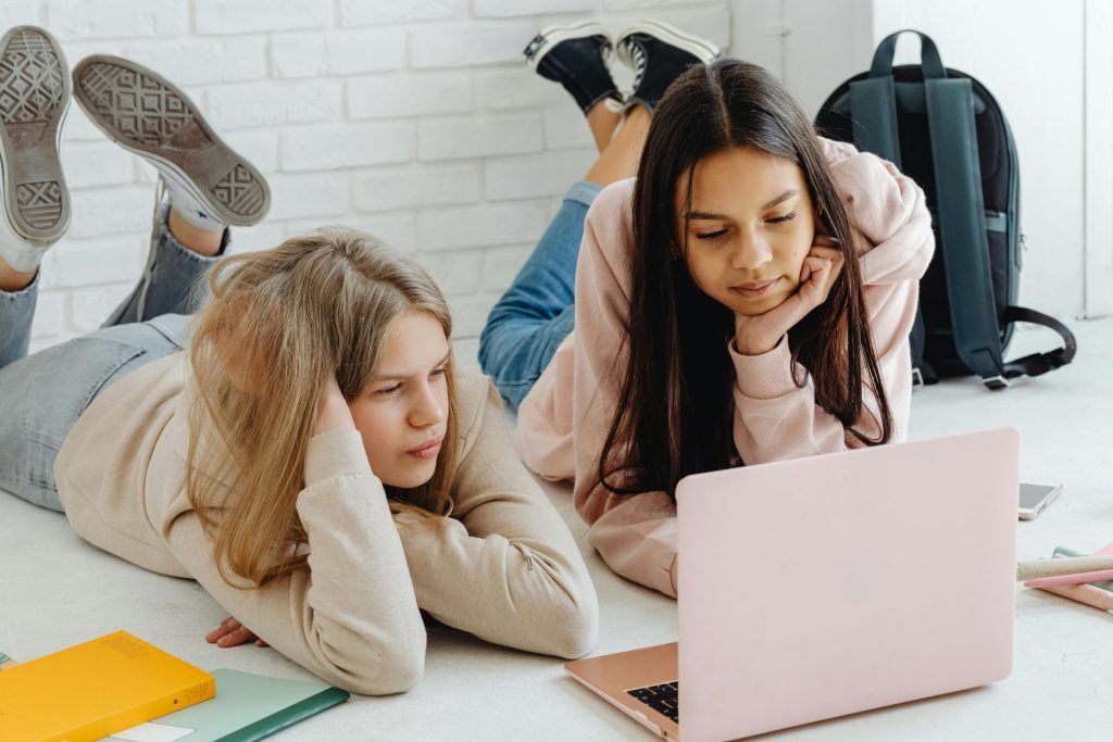 Two teenage girls lying on the floor, propped on their elbows, looking at a pink laptop. School supplies and a backpack are nearby, and the scene appears relaxed and focused.