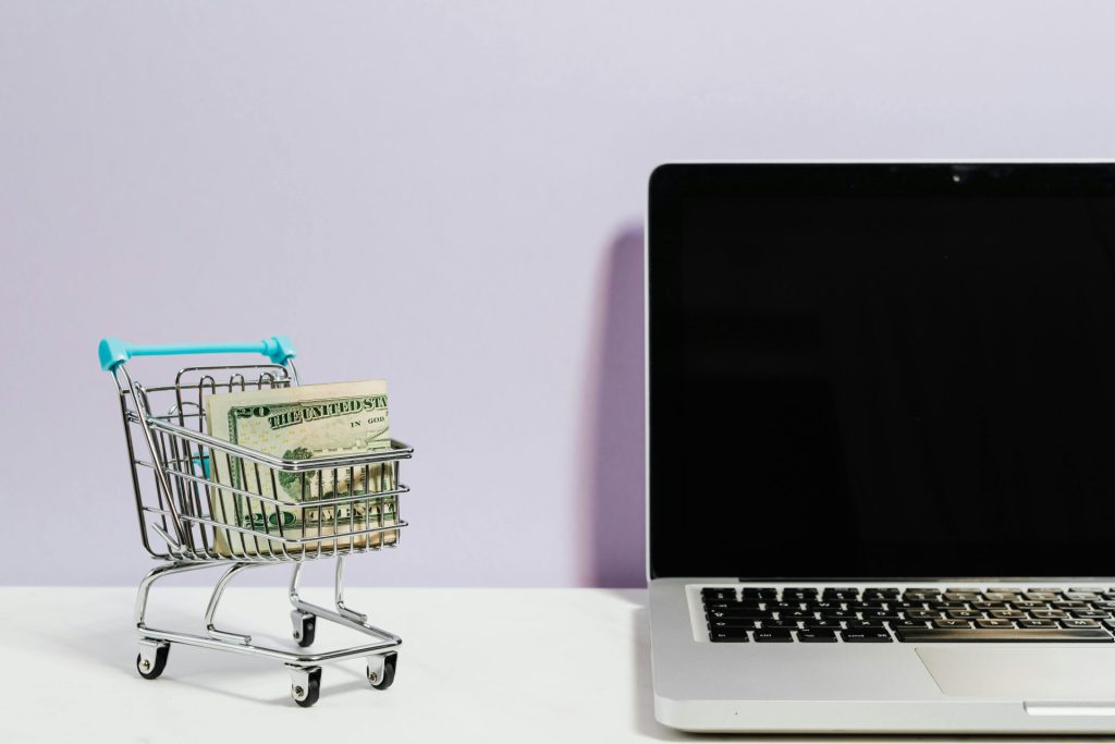 A small shopping cart filled with US dollar bills is placed next to an open laptop with a blank screen on a white surface against a light purple background.