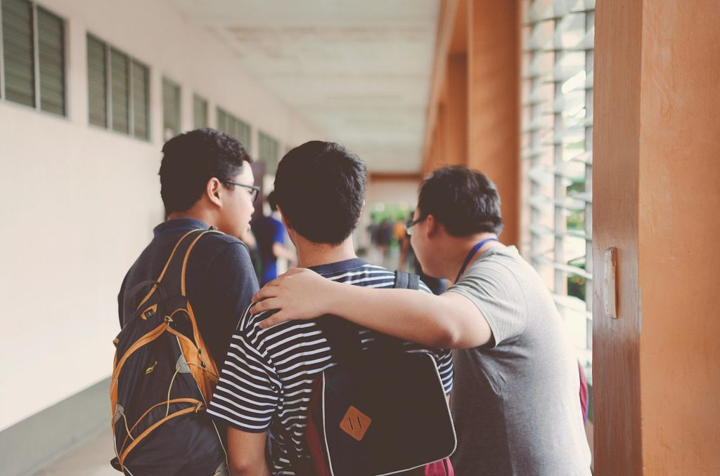 Three students with backpacks walk down a hallway. One student has his arm around another as they talk. The hallway has orange pillars and large windows with bars on one side.