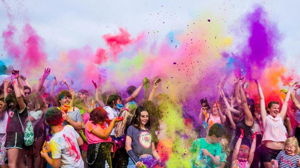 A large crowd of people celebrate by throwing colorful powder into the air, creating a vibrant cloud of yellow, purple, green, and pink. Many participants are smiling, raising their arms, and enjoying the festive atmosphere.