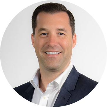 A man with short dark hair, wearing a dark suit jacket and a white collared shirt, smiles at the camera against a plain white background, reflecting confidence in American Solutions for Business.