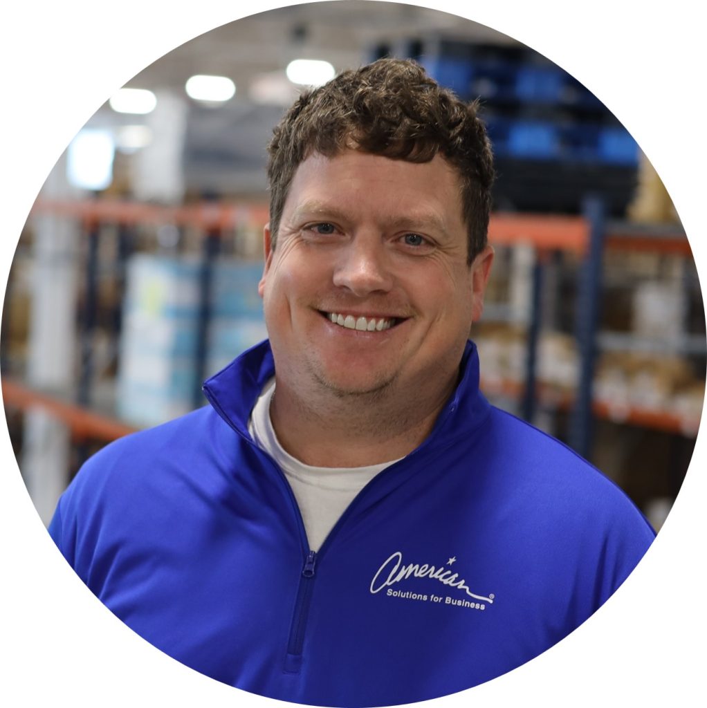 A smiling man with short brown hair wears a blue "American Solutions for Business" jacket, standing in a warehouse with shelves in the background, representing seamless integration of platforms like PeopleSoft and SAGE.