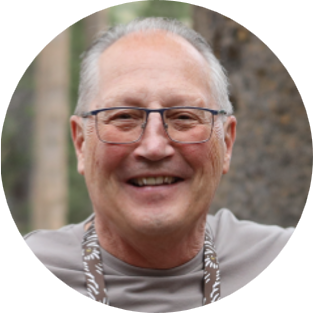 An older man with gray hair and glasses smiles warmly outdoors. Wearing a gray shirt and a patterned lanyard, he appears at an American Solutions for Business event, with trees visible in the blurred background.