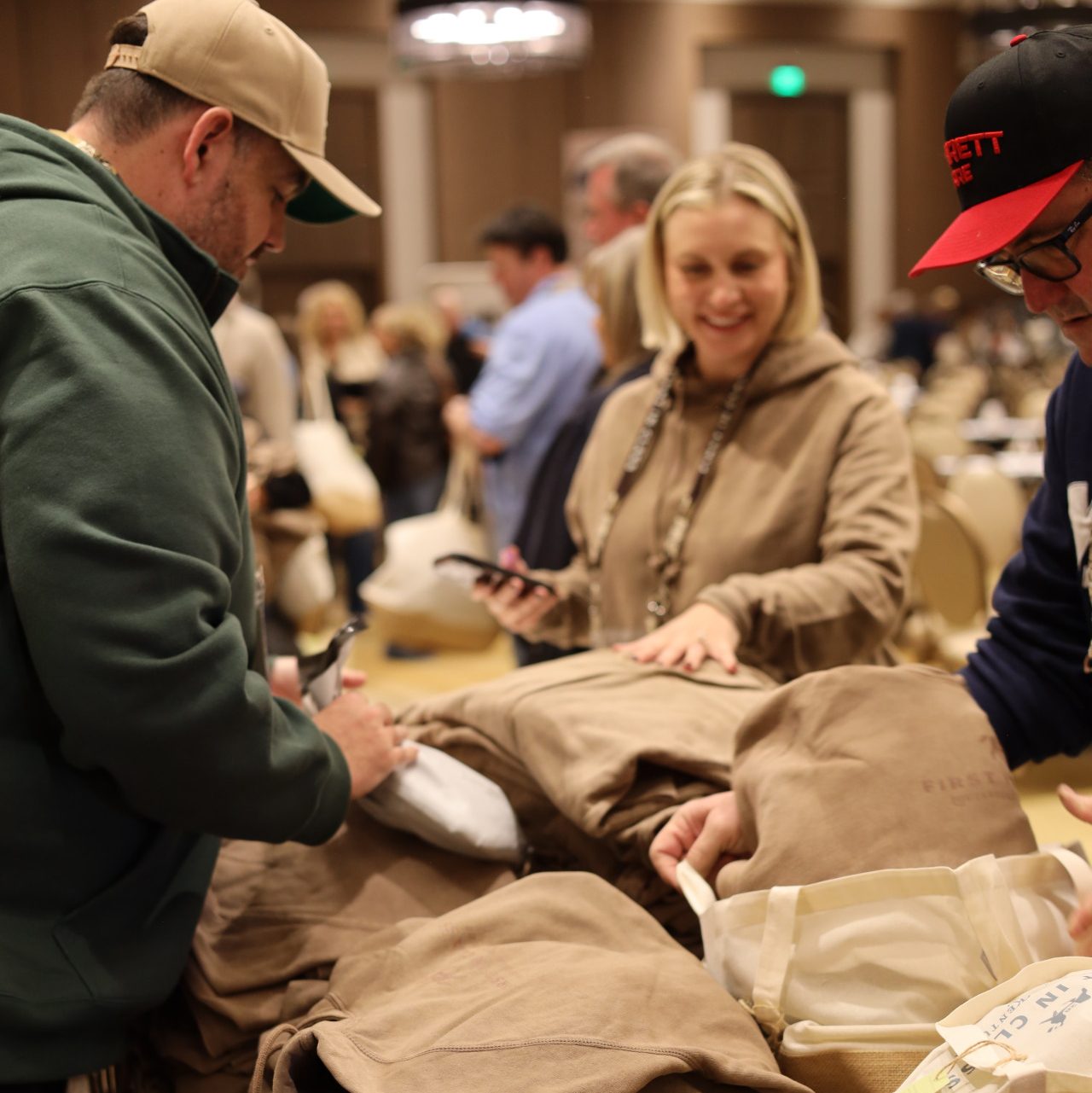 People gather around a table covered with folded brown shirts and tote bags at the National Sales Conference in Louisville, Kentucky. A woman smiles while holding her phone as others browse the ASB-branded items at this busy indoor event.