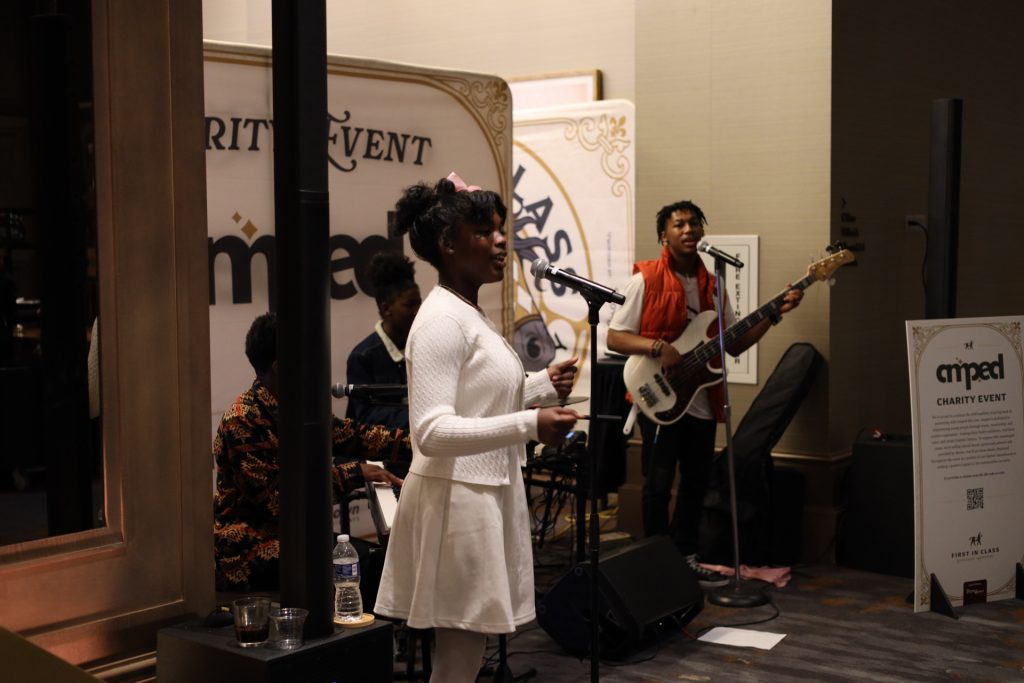 A young woman in a white dress sings into a microphone while a band performs behind her, featuring a bassist and keyboardist, at an indoor ASB charity event. A sign and event backdrop are visible in the background.