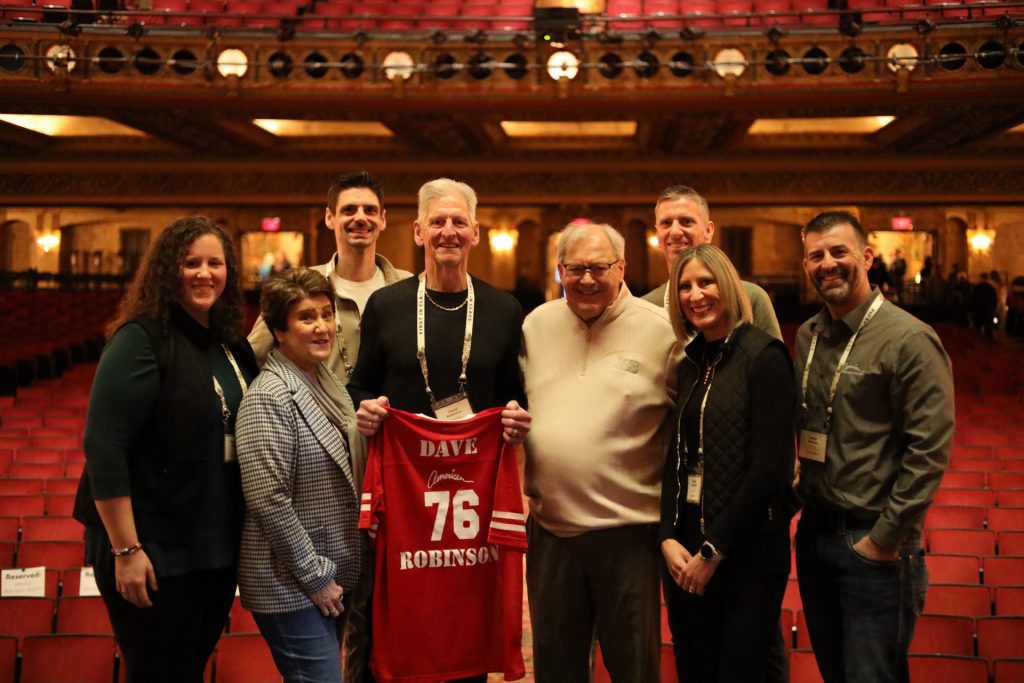 A group of eight people smile for a photo at the National Sales Conference in a Louisville Kentucky theater, with one person holding up a red "DAVE 76 ROBINSON" jersey. The ornate décor and theater seats are visible in the background.