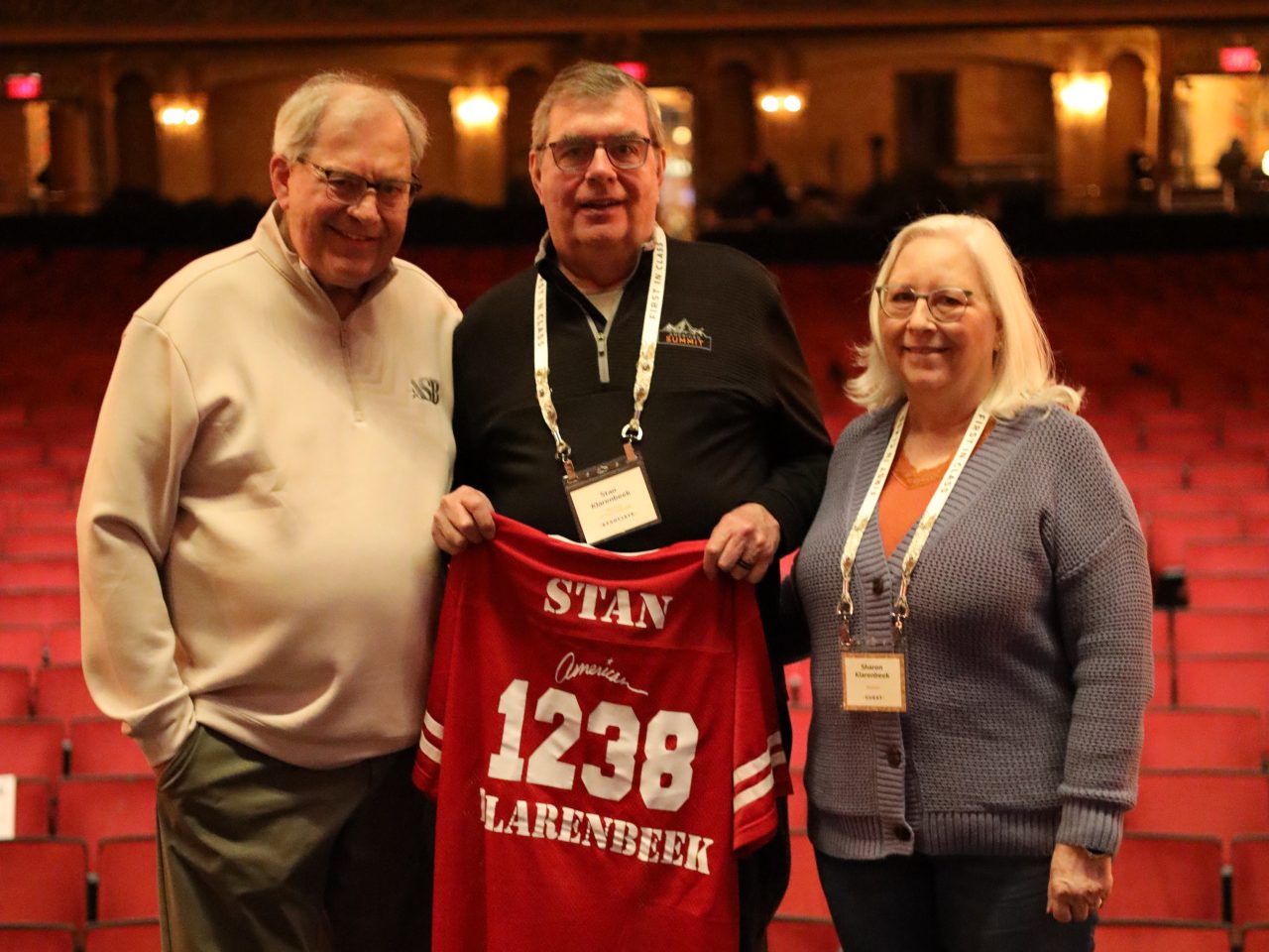 Three adults stand on a theater stage in Louisville, Kentucky, smiling at the camera. The man in the center holds a red hockey jersey with "STAN 1238 CLARENBEEK" on it. The ASB national sales conference's empty red seats are visible behind them.
