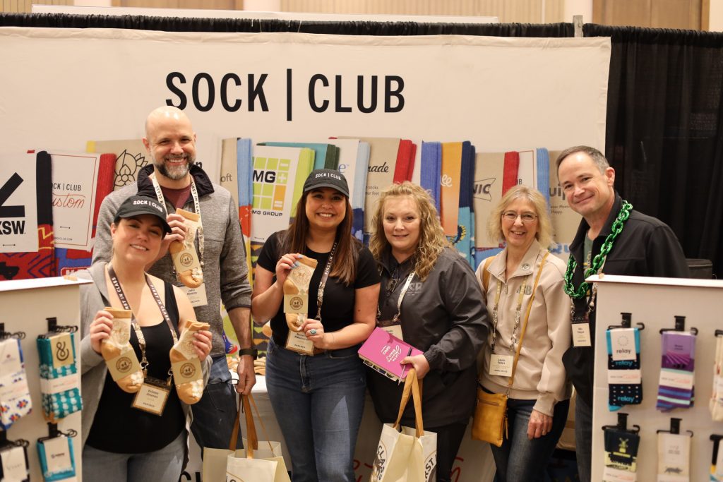 Six people stand smiling in front of a “Sock Club” booth at the National Sales Conference in Louisville, Kentucky, holding socks and tote bags. Colorful socks decorate the booth as the ASB group appears happy and engaged at the event.