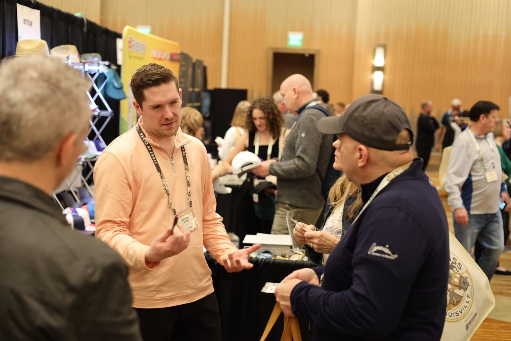 At the National Sales Conference in Louisville, Kentucky, a man in a peach shirt talks to a man in a navy cap as people network and ASB booths fill the busy indoor event space.