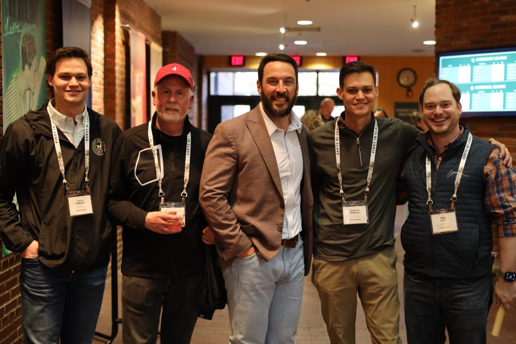 Five men wearing name badges stand together smiling indoors, in a warmly lit space with brick walls and digital screens—captured at the ASB National Sales Conference in Louisville, Kentucky.