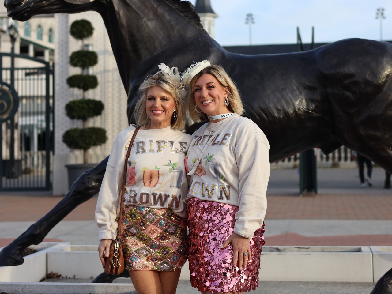 Two women stand smiling in front of a horse statue at Churchill Downs in Louisville, Kentucky. Both wear matching “Triple Crown” sweatshirts, decorative headpieces, and sequined skirts, posing for a celebratory photo at the racetrack entrance.