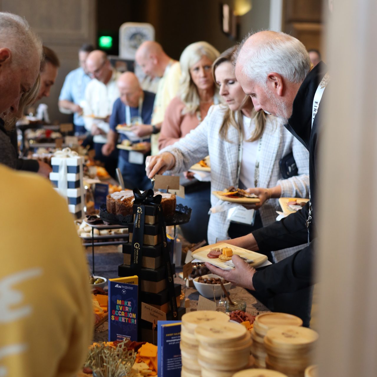 A group of people serves themselves food from a buffet table filled with a variety of dishes at the busy, lively National Sales Conference hosted by ASB in Louisville, Kentucky. Many guests hold plates and reach for food as they enjoy the indoor event.