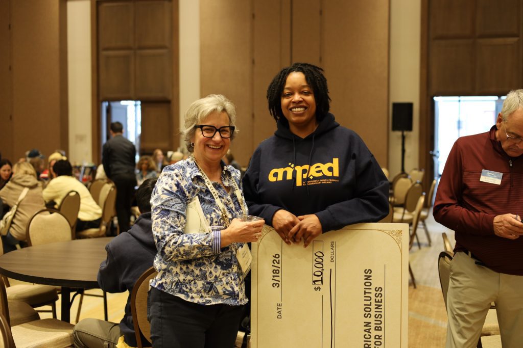 Two women smile at the camera while holding a large $10,000 check for charity at an indoor event. Attendees of the National Sales Conference are seated at round tables in the background. One woman wears glasses and a floral shirt, the other a dark hoodie.