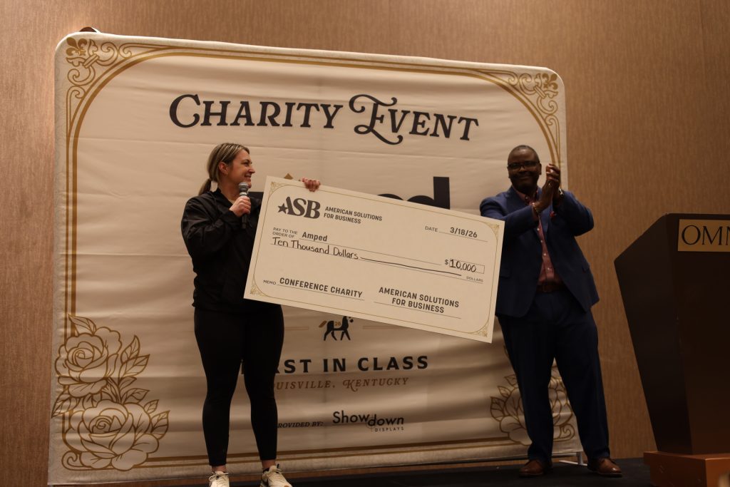 A woman holding a $10,000 check from ASB for the charity Amped stands on stage with a microphone at the National Sales Conference, while a man next to her claps.