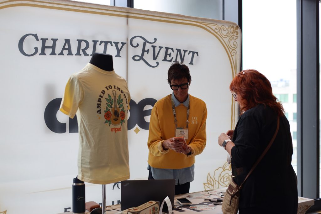Two people stand at a booth with a "Charity Event" sign, displaying a yellow T-shirt and merchandise. One assists the other looking at items, highlighting ASB's commitment to charity. Large windows are in the background.