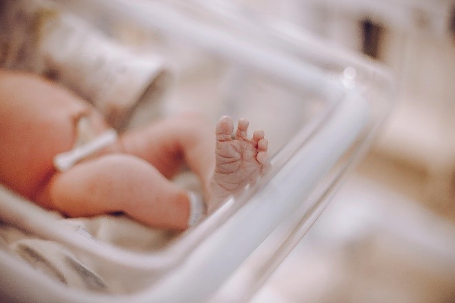 A close-up of a newborn baby lying in an incubator, showing a tiny foot and hand. The baby has medical tubes attached, indicating care in a neonatal unit—capturing early life events often supported by financial institutions. The background is softly blurred.