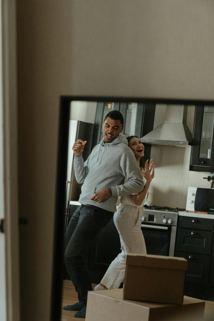 A couple is dancing and laughing together in a kitchen, seen through a doorway with cardboard moving boxes in the foreground—a joyful moment celebrating life events. The kitchen has dark cabinets and stainless steel appliances.