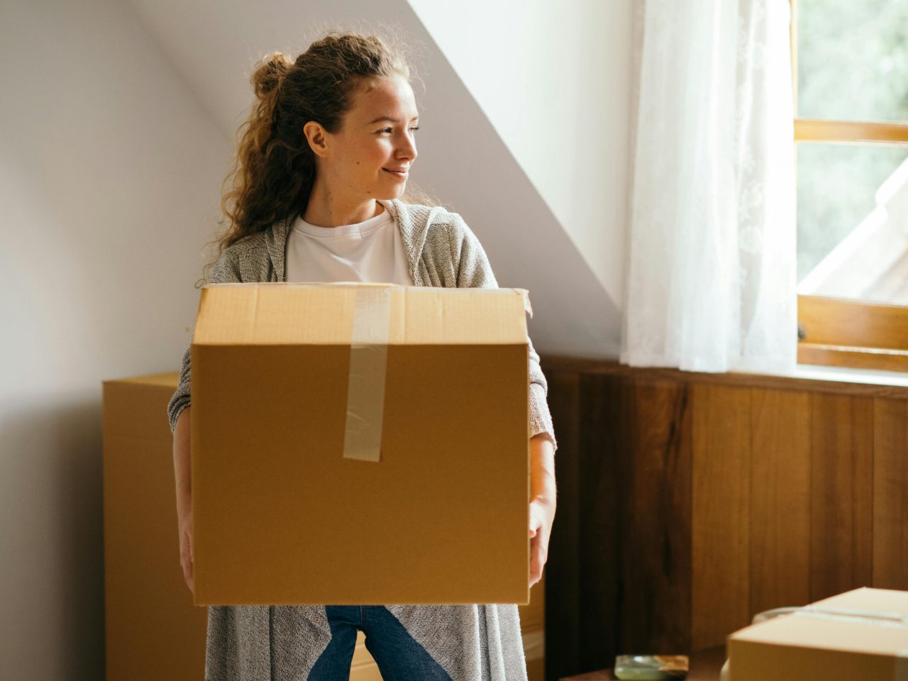 A woman stands in a bedroom holding a large cardboard box, smiling and looking toward a window. Surrounded by packed boxes and sunlight, she embraces one of life events that often create marketing opportunities for financial institutions.