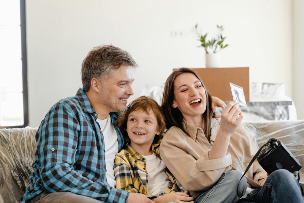 A smiling couple sits on a couch with their young son between them. The woman holds a photo and laughs while the man and child smile, enjoying a happy moment—one of those life events cherished in any cozy home setting.
