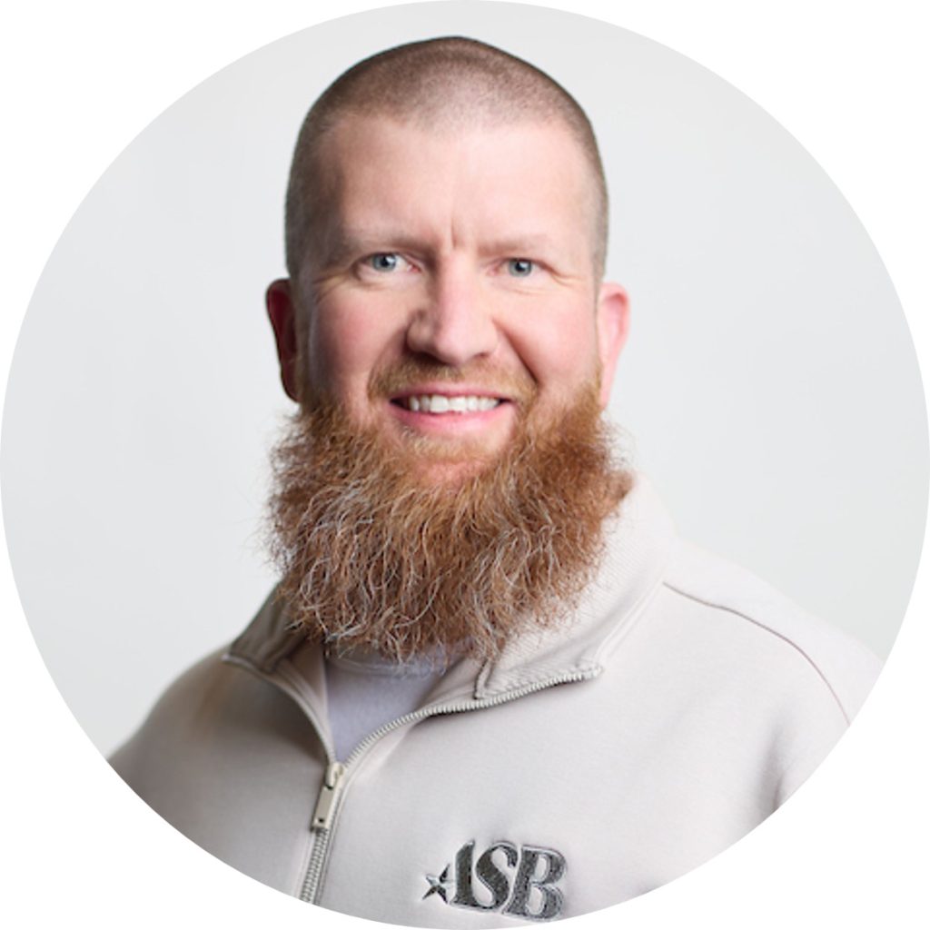 A smiling man with a reddish-brown beard and short hair, Nate Disrud, Senior Director of Technology at American Solutions for Business, wears a light-colored zip-up jacket with "ASB" embroidered on it, in front of a plain white background.