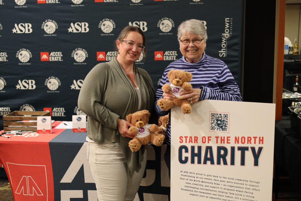Two smiling women hold teddy bears in front of a charity display for Star of the North Maternity Home at the Accelerate Event. The sign features a QR code, with an ASB-branded backdrop and event table visible in the background.