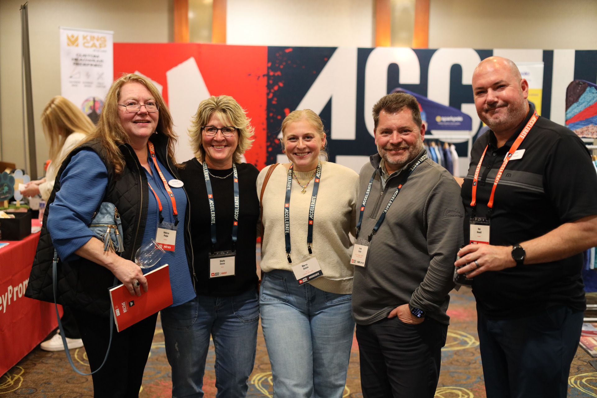 Five smiling adults, three women and two men, stand together indoors at the ASB Accelerate Event in Duluth, MN. They wear name badges and casual clothing, with a colorful display background behind them at this 2026 conference.