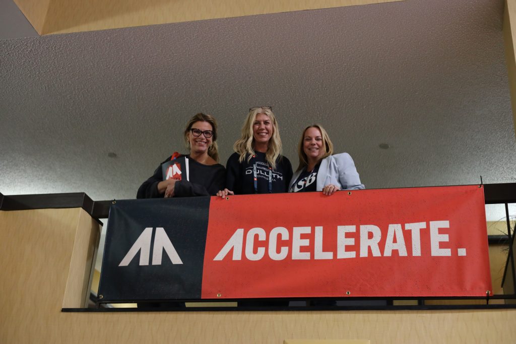 Three women stand on a balcony behind a red and black "ACCELERATE" banner with a stylized "AA" logo at the ASB Accelerate Event in Duluth, MN. They are smiling and looking at the camera indoors, capturing the energy of 2026.