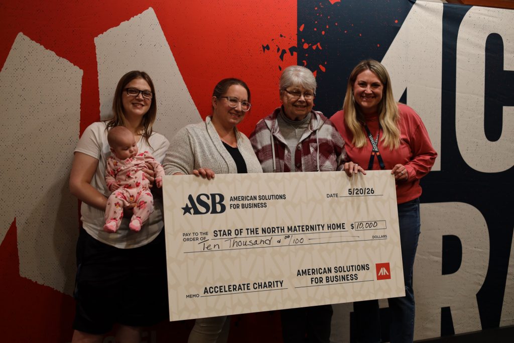 Four smiling women, one holding a baby, pose in front of a wall with an oversized $10,000 check for Star of the North Maternity Home from ASB, presented at the Accelerate Event.