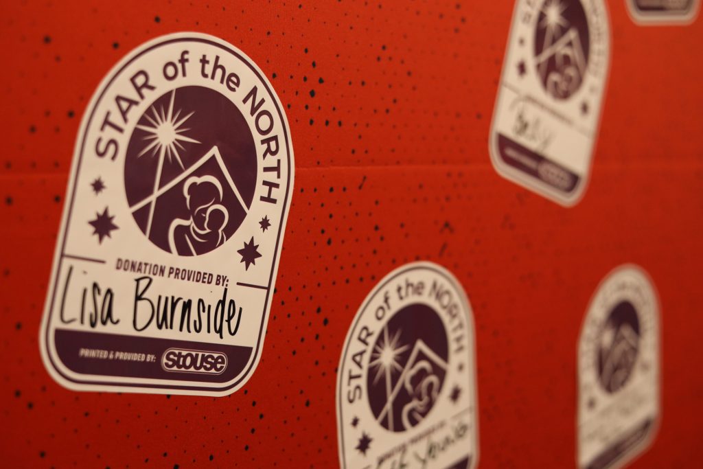 White "Star of the North" stickers with a stylized family design are placed on a red speckled background. One sticker reads "Donation provided by: Lisa Burnside" in handwritten text, supporting Star of the North Maternity Home's Accelerate Event.