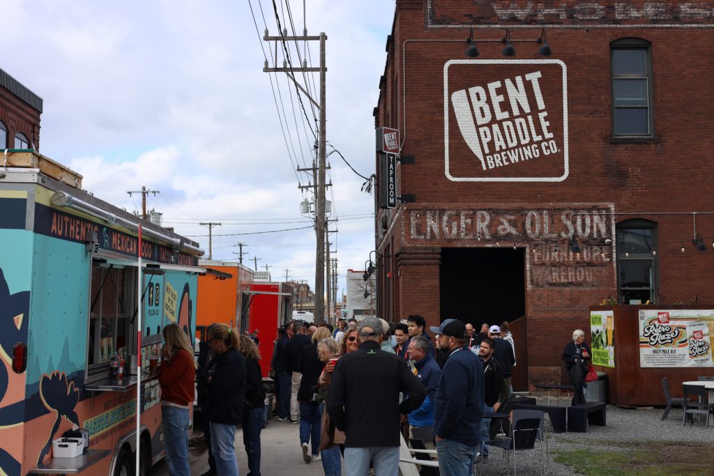 A crowd gathers near food trucks outside Bent Paddle Brewing Co., a brick building with painted signage in Duluth MN. People stand in line and chat on a cloudy day during the lively ASB Accelerate Event in this urban setting.